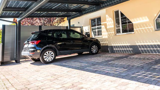 Schwarzes Auto parkt unter Carport neben gelbem Wohnhaus mit gepflasterter Einfahrt. HANSEL.kraus metalltechnik gmbh