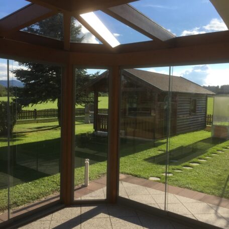Wintergarten mit Glaswänden und Blick auf grünen Garten und Holzhütte bei Sonnenschein. HANSEL.kraus metalltechnik gmbh