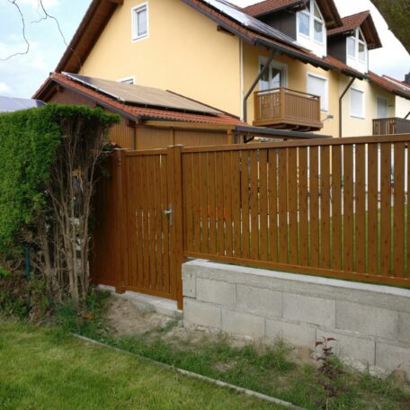 Holzzaun mit Tür vor gelbem Haus, Rasen und bepflanzter Hecke im Vordergrund. de HANSEL.kraus metalltechnik gmbh