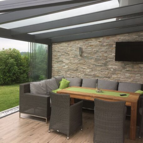 Gemütliche Terrasse mit Glasdach, Holztisch, grauen Sitzmöbeln und grünem Garten im Hintergrund. HANSEL.kraus metalltechnik gmbh