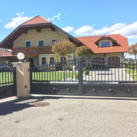 Großes Einfamilienhaus mit rotem Dach, Garten und geschlossenem Metalltor bei Sonnenschein. HANSEL.kraus metalltechnik gmbh
