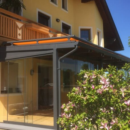 Wintergarten mit Glasdach an gelbem Einfamilienhaus, davor blühender Strauch und blauer Himmel. HANSEL.kraus metalltechnik gmbh