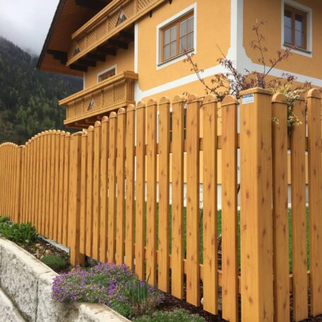 Holzzaun vor gelbem Haus mit Blumenkasten und Gebirge im Hintergrund. HANSEL.kraus metalltechnik gmbh