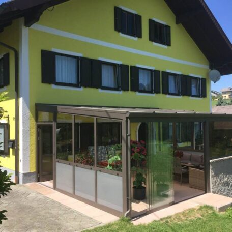 Wintergarten an gelbem Einfamilienhaus mit Glasdach, Garten und Sitzbereich bei Sonnenschein. HANSEL.kraus metalltechnik gmbh