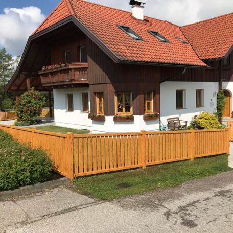 Einfamilienhaus mit rotem Dach, Holzbalkon und Garten, von Holzzaun umgeben, bei Sonnenschein. HANSEL.kraus metalltechnik gmbh