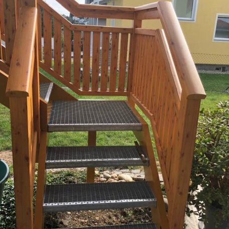 Holztreppe mit Metallstufen führt in einen Garten, gelbes Haus im Hintergrund. HANSEL.kraus metalltechnik gmbh