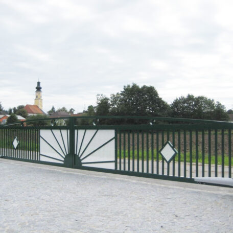 Tor mit modernem Muster, Kiesweg davor, Dorf und Kirche im Hintergrund, bewölkter Himmel. HANSEL.kraus metalltechnik gmbh