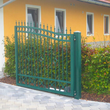 Grünes Einfahrtstor aus Metall vor gelbem Haus mit Pflasterweg und Hecke. de HANSEL.kraus metalltechnik gmbh