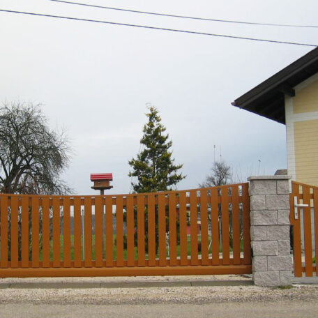 Holzzaun mit Steinpfosten vor einem Einfamilienhaus, dahinter Garten mit Baum und Vogelhaus. de HANSEL.kraus metalltechnik gmbh