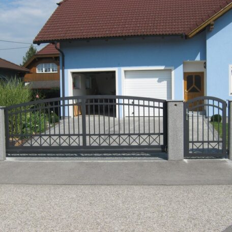 Metallzaun mit Einfahrtstor vor einem blauen Einfamilienhaus und offener Garage. HANSEL.kraus metalltechnik gmbh