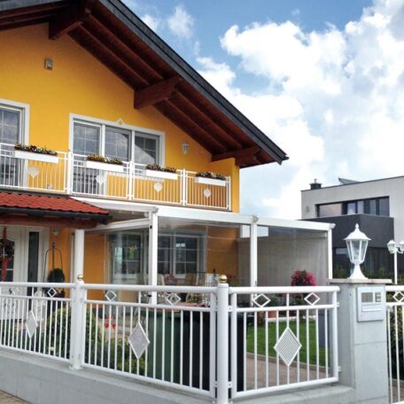 Zweistöckiges gelbes Haus mit weißem Zaun und Balkon, moderner Neubau im Hintergrund. HANSEL.kraus metalltechnik gmbh