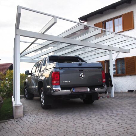 Pickup unter modernem Glas-Carport neben Wohnhaus, gepflasterte Einfahrt, Tageslicht. de HANSEL.kraus metalltechnik gmbh