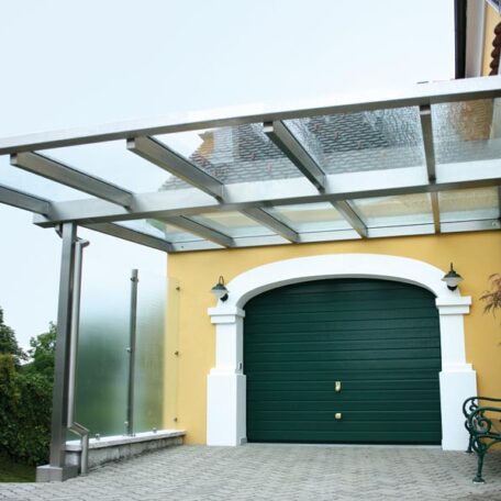 Carport mit transparentem Dach vor einer gelben Garage mit grünem Tor. HANSEL.kraus metalltechnik gmbh
