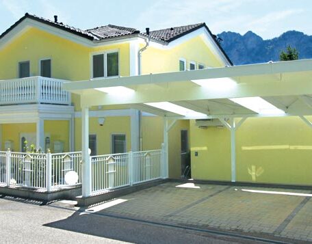 Zweistöckiges gelbes Haus mit überdachtem Carport und Bergen im Hintergrund. HANSEL.kraus metalltechnik gmbh