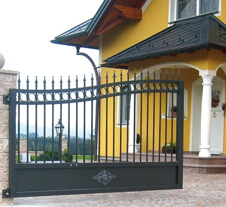 Schwarzes schmiedeeisernes Einfahrtstor vor gelbem Einfamilienhaus mit Säulen und überdachtem Eingangsbereich. HANSEL.kraus metalltechnik gmbh