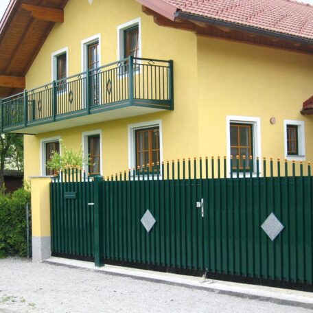 Gelbes Haus mit grünem Zaun, Balkon und mehreren Fenstern, Einfahrtstor geschlossen. HANSEL.kraus metalltechnik gmbh
