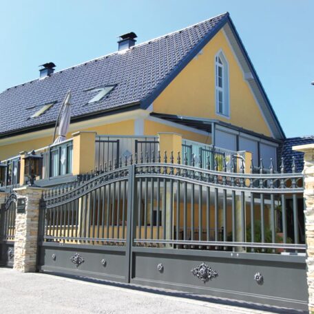 Modernes gelbes Einfamilienhaus mit großem Metallzaun und Einfahrtstor bei Sonnenschein. HANSEL.kraus metalltechnik gmbh