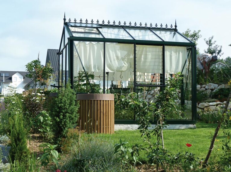 Glashaus im Garten mit Pflanzen und Bäumen, dekorative Vorhänge im Inneren, Tageslicht. HANSEL.kraus metalltechnik gmbh