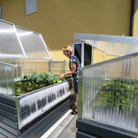 Eine Person pflegt Pflanzen in einem Hochbeet-Gewächshaus auf einer sonnigen Terrasse. de HANSEL.kraus metalltechnik gmbh