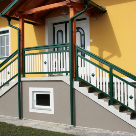 Hauseingang mit grüner Treppe und Geländer, gelbe Fassade, kleines Kellerfenster darunter. de HANSEL.kraus metalltechnik gmbh