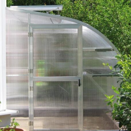 Gewächshaus mit transparenter Tür steht im Garten, umgeben von grünen Pflanzen. de HANSEL.kraus metalltechnik gmbh