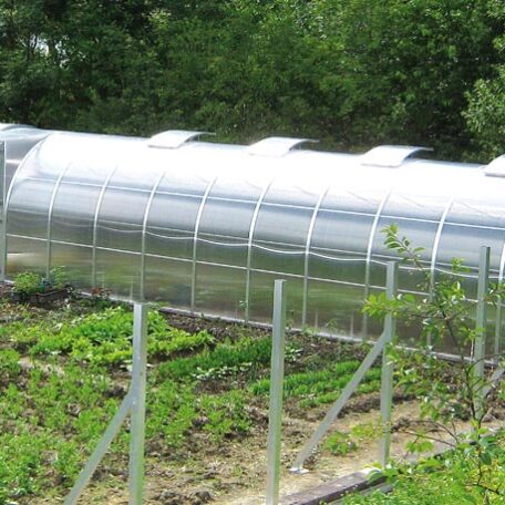 Ein längliches Gewächshaus steht in einem Garten mit bepflanzten Gemüsebeeten und grüner Umgebung. HANSEL.kraus metalltechnik gmbh