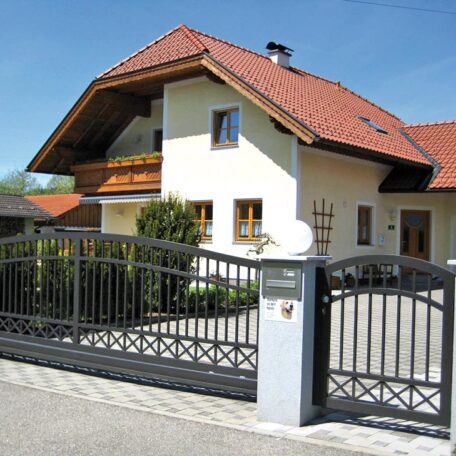 Modernes Einfamilienhaus mit rotem Ziegeldach hinter dekorativem Metallzaun bei sonnigem Wetter. HANSEL.kraus metalltechnik gmbh