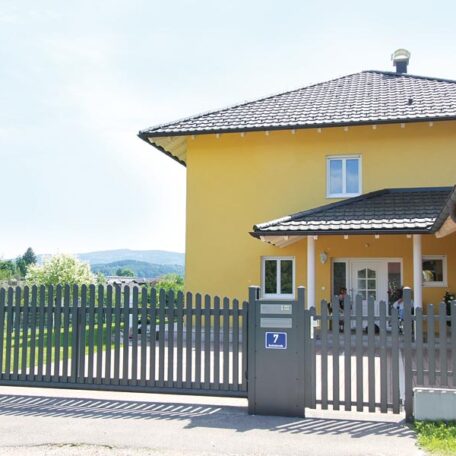 Gelbes Einfamilienhaus mit Garten, Holzzaun und Terrasse an sonnigem Tag in ruhiger Wohngegend. HANSEL.kraus metalltechnik gmbh