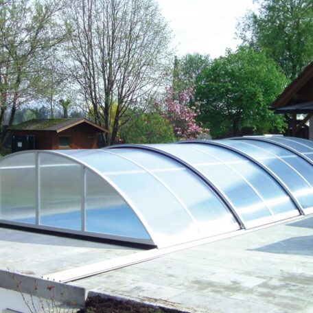 Gewölbte Poolüberdachung aus Glas neben Garten mit Bäumen und Holzhäusern. HANSEL.kraus metalltechnik gmbh
