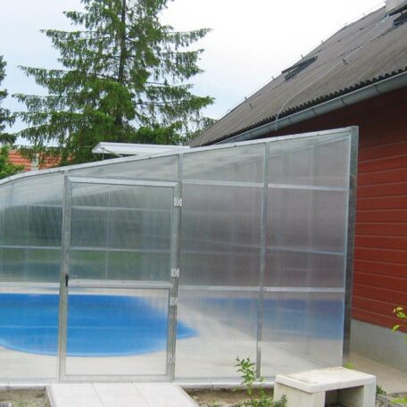 Überdachtes Schwimmbecken im Garten neben einem roten Wohnhaus bei Tageslicht. HANSEL.kraus metalltechnik gmbh