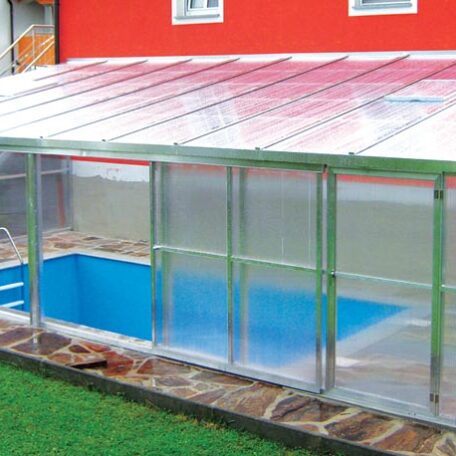 Überdachter Pool mit glasklarer Poolüberdachung neben einem roten Wohnhaus auf Steinboden. HANSEL.kraus metalltechnik gmbh