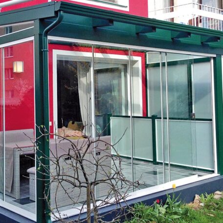Moderne Terrassenüberdachung aus Glas an rotem Wohnhaus mit Garten, Tageslichtszene. HANSEL.kraus metalltechnik gmbh