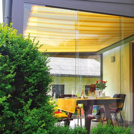 Wintergarten mit gelber Markise, Esstisch und Stühlen, Blick durch große Glasfront, grüner Busch davor. HANSEL.kraus metalltechnik gmbh