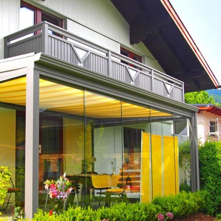 Eine moderne Terrassenüberdachung mit Glaswänden an einem Einfamilienhaus, umgeben von Garten und Balkon. HANSEL.kraus metalltechnik gmbh
