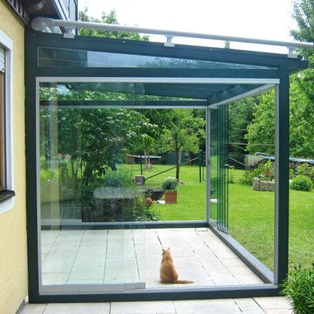 Katze sitzt in verglaster Terrassenüberdachung, dahinter grüner Garten mit Bäumen. HANSEL.kraus metalltechnik gmbh
