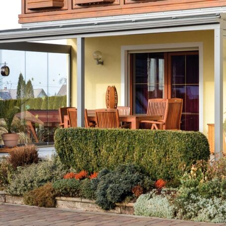 Eine überdachte Terrasse mit Holzmöbeln und gepflegtem Vorgarten vor einem gelben Haus. de HANSEL.kraus metalltechnik gmbh