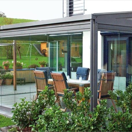 Wintergarten mit Glaswänden und Holzmöbeln, angrenzend an einen grünen Garten, bei Tageslicht. HANSEL.kraus metalltechnik gmbh