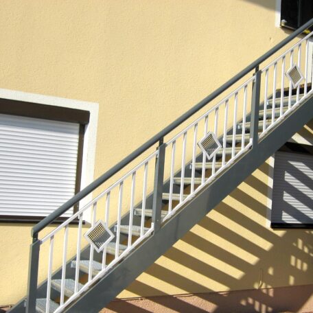 Außentreppe mit Geländer wirft Schatten auf gelbe Hauswand mit geschlossenem Fenster. HANSEL.kraus metalltechnik gmbh