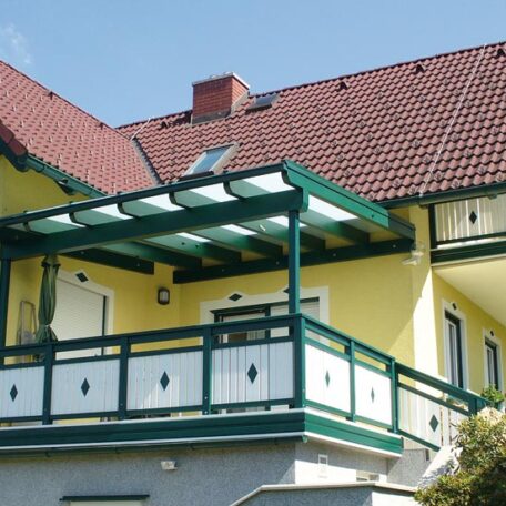 Überdachter Balkon mit grünem Geländer an gelbem Wohnhaus unter blauem Himmel. HANSEL.kraus metalltechnik gmbh