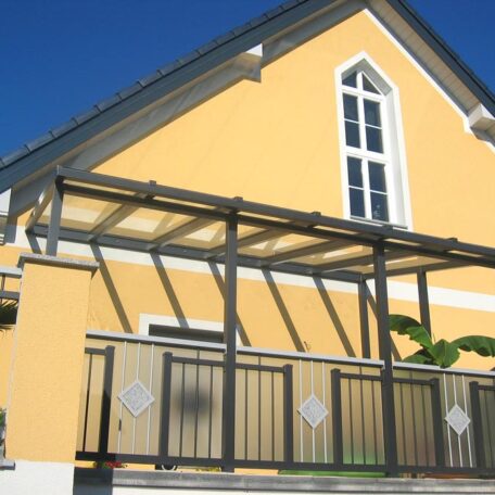 Modernes gelbes Haus mit Wintergarten-Anbau und dekorativem Metallzaun, blauer Himmel. de HANSEL.kraus metalltechnik gmbh