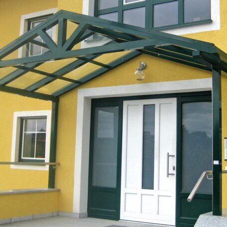 Grünes Vordach aus Glas und Metall über weißer Haustür an gelbem Gebäude. HANSEL.kraus metalltechnik gmbh