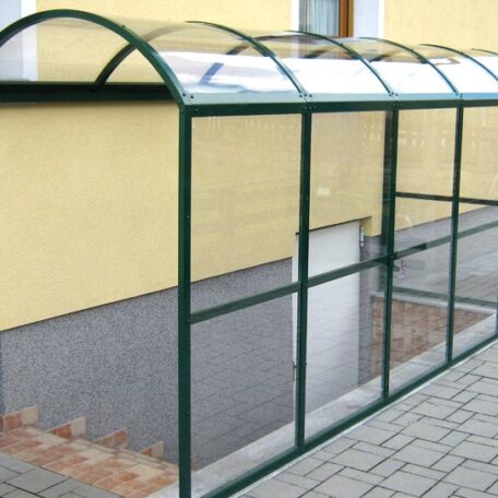 Überdachter Fahrradunterstand mit grünem Gestell vor gelber Hauswand auf gepflastertem Boden. HANSEL.kraus metalltechnik gmbh