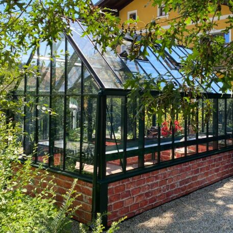 Modernes Gewächshaus aus Glas und Metall steht im sonnigen Garten vor gelbem Wohnhaus. de HANSEL.kraus metalltechnik gmbh