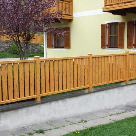 Holzzaun auf Betonmauer vor gelbem Wohnhaus mit offenen braunen Fensterläden und grünem Garten. HANSEL.kraus metalltechnik gmbh
