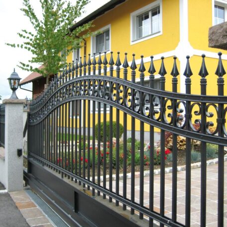 Schwarzes schmiedeeisernes Hoftor vor gelbem Wohnhaus mit gepflegtem Vorgarten und Laterne. de HANSEL.kraus metalltechnik gmbh