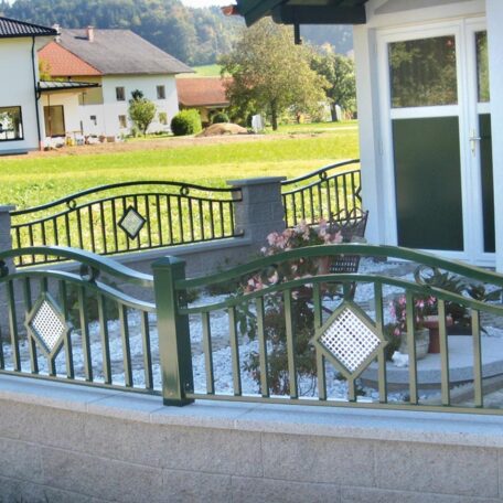 Metallzaun mit dekorativen Elementen vor einem Haus, im Hintergrund grüne Wiese und weitere Gebäude. HANSEL.kraus metalltechnik gmbh