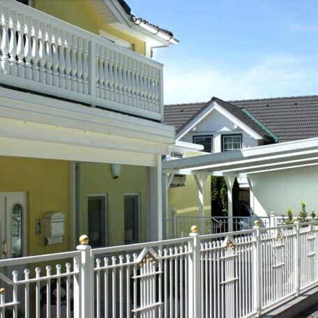 Gelbes Einfamilienhaus mit weißem Balkon, Zaun und überdachtem Eingangsbereich bei Tageslicht. HANSEL.kraus metalltechnik gmbh