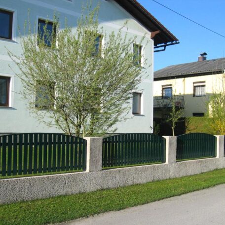 Zweistöckiges Haus mit grünem Zaun, Vorgarten und Baum an sonniger Straße. de HANSEL.kraus metalltechnik gmbh