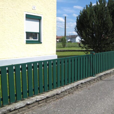 Ein grüner Holzzaun steht vor einem gelben Haus mit geschlossenem Fenster und angrenzendem Gehweg. HANSEL.kraus metalltechnik gmbh