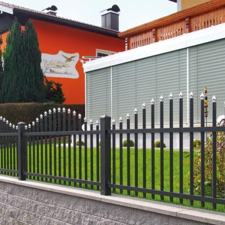 Moderner Metallzaun vor einem orangefarbenen Haus mit gepflegtem grünen Vorgarten. HANSEL.kraus metalltechnik gmbh